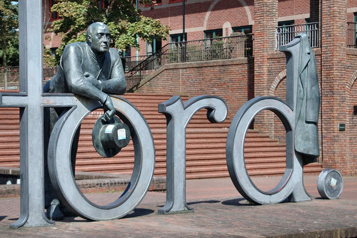 Statue of man leaning on 'Telford' sign in urban setting.