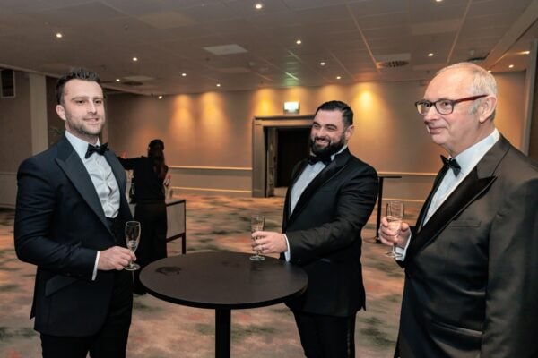 Three men in formal tuxedos at a professional awards event, holding champagne glasses, engaging in conversation in an elegant, well-lit venue.