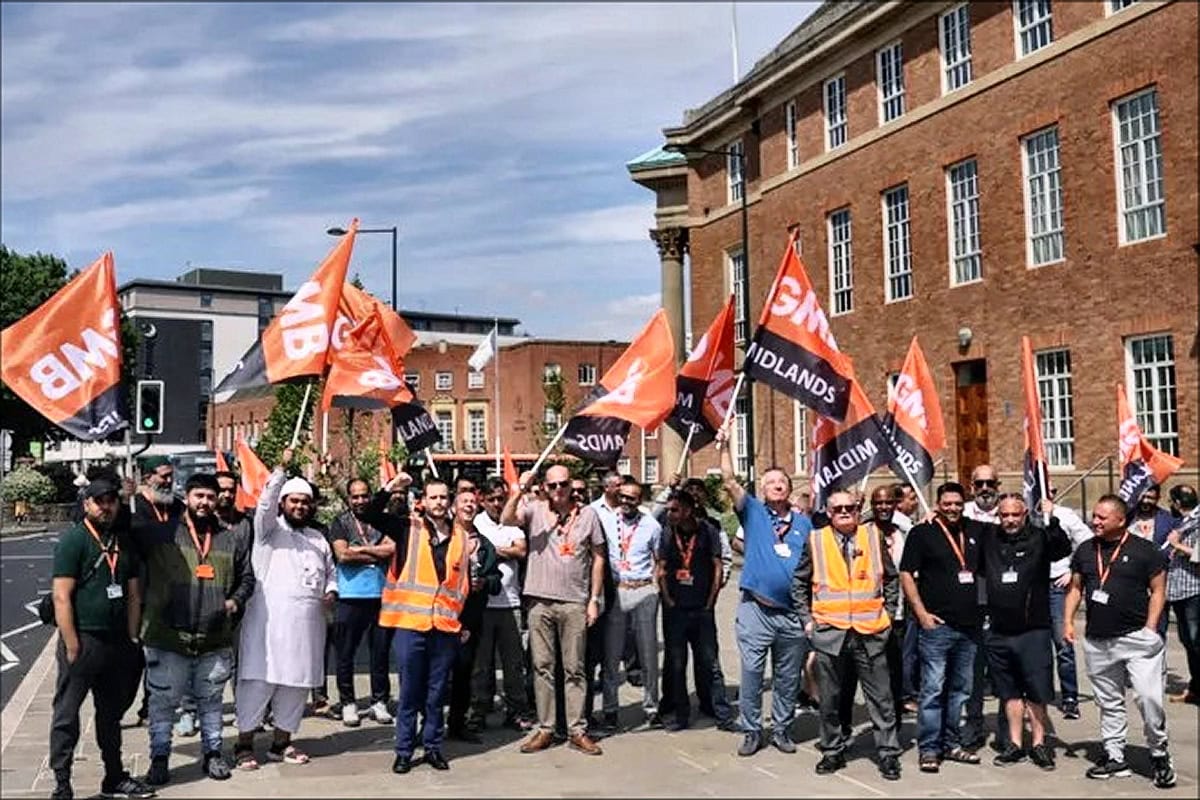 Protesters holding orange flags and banners, advocating for the extension of the Private Hire Vehicle age limit, in front of a historic brick building in Derby.