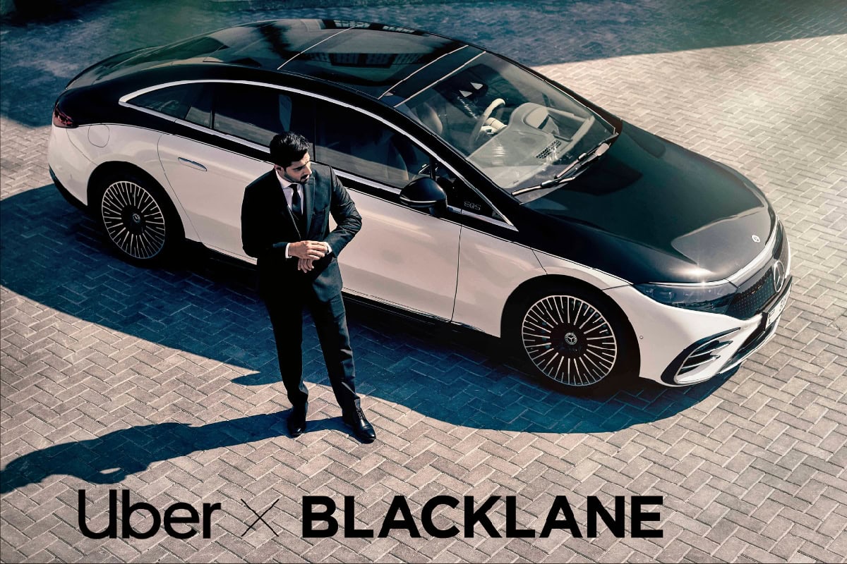 An image of a professional chauffeur standing beside a luxury black and white Uber vehicle, highlighting the recent acquisition of Blacklane by Uber to strengthen its position in the chauffeur sector.