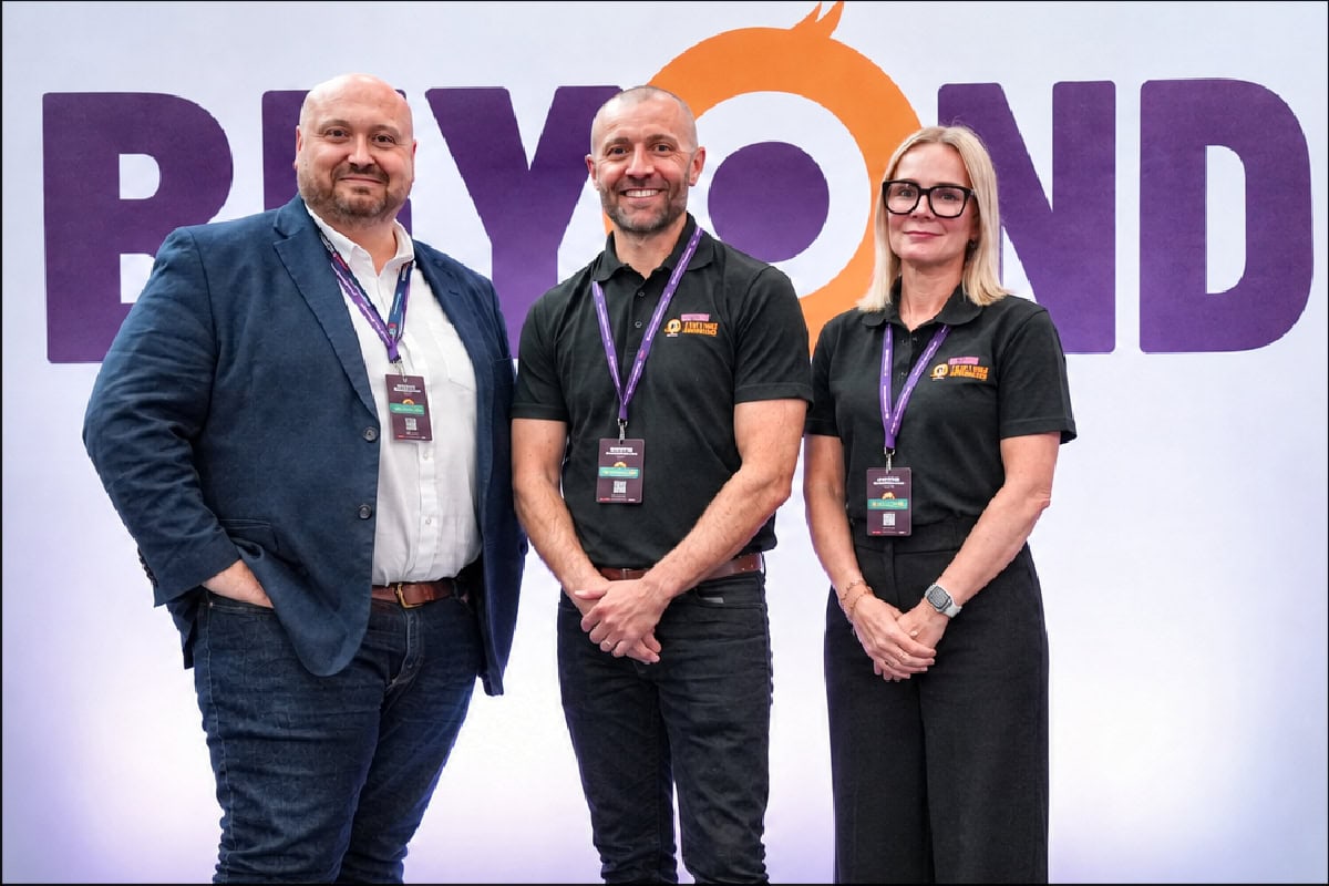 Three professional individuals at a conference, standing in front of a 'BEYOND' banner, representing the Coach Hire Comparison industry.