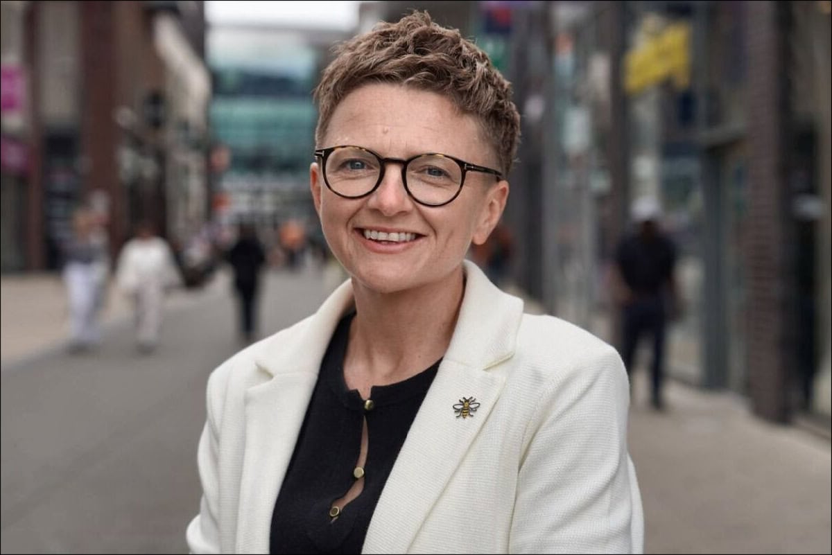 Smiling woman with short hair and glasses standing on a busy city street in Greater Manchester.