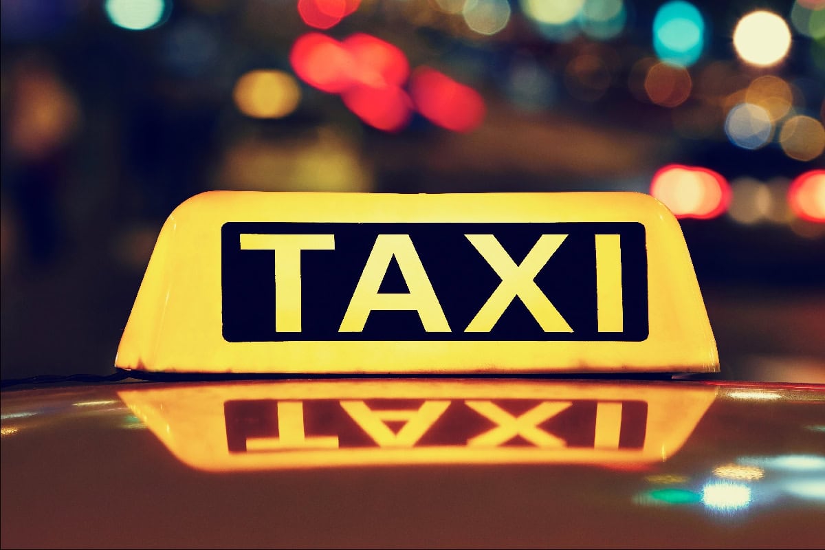 Bright yellow taxi sign with black 'TAXI' lettering on a vehicle at night, with blurred city lights in the background.